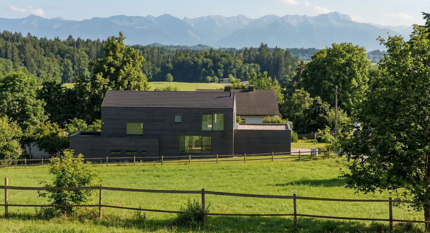 Modern dark gray house with large windows, surrounded by green grass, trees, and mountains in the background.
