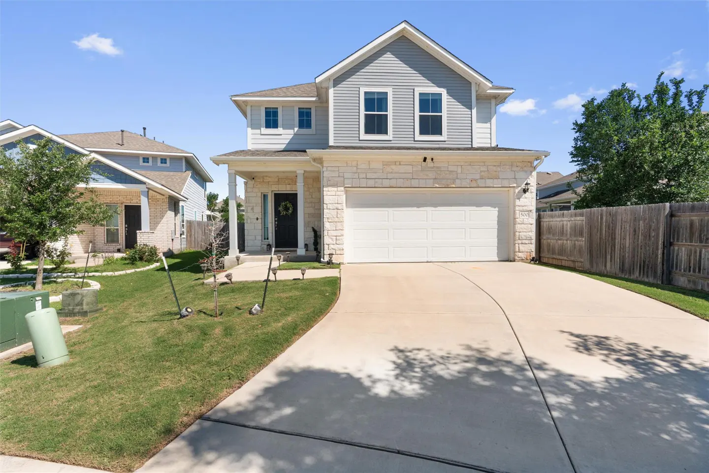 Two-story house with gray siding, stone accents, white trim, and a two-car garage on a sunny day.