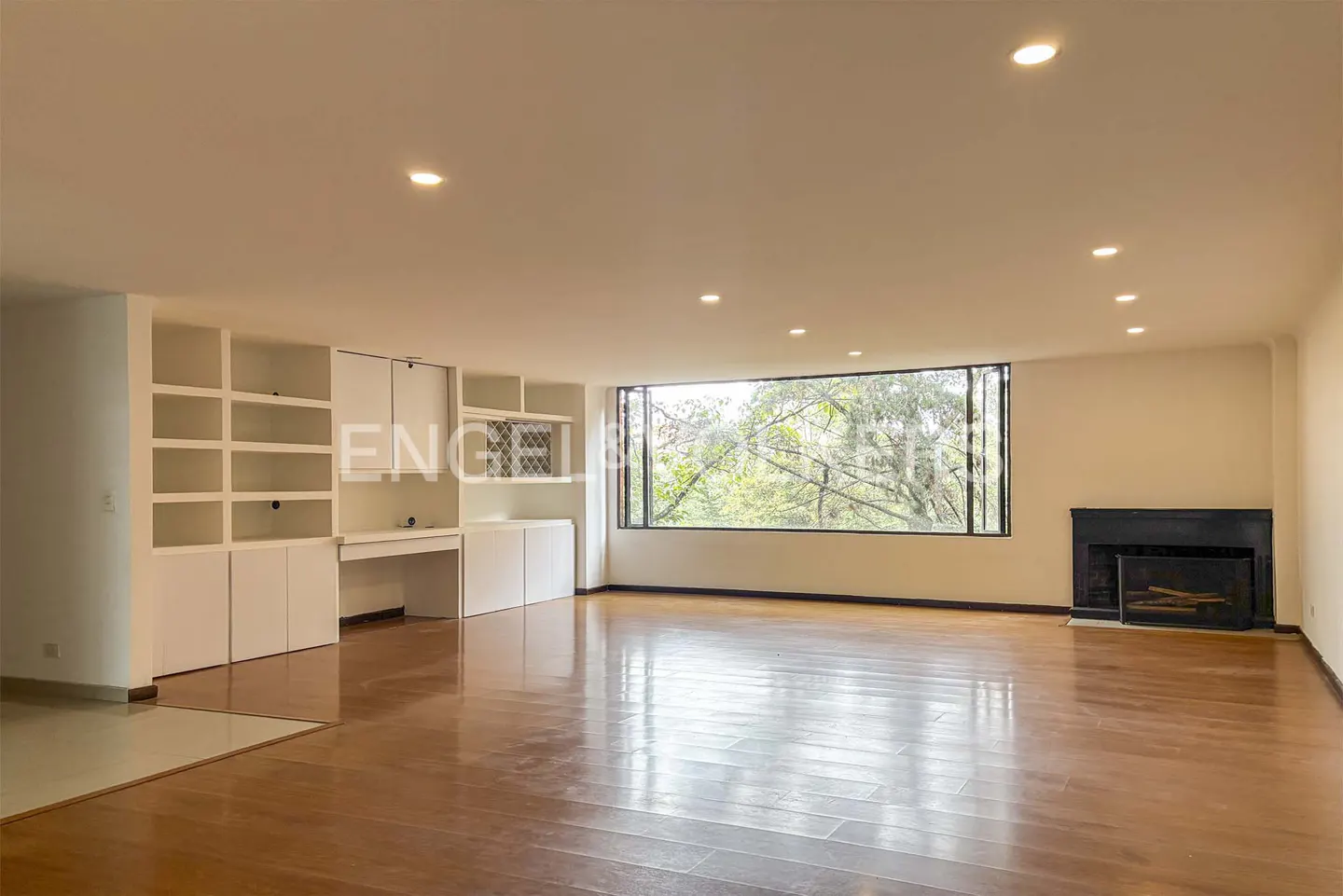 Spacious living room with hardwood floors, white built-in shelving, a large window with tree views, and a black fireplace.