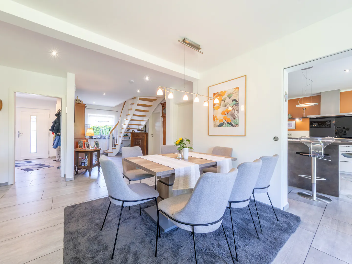 Dining area with a wooden table, six grey chairs on a rug, modern lighting, and a view of the staircase and kitchen.