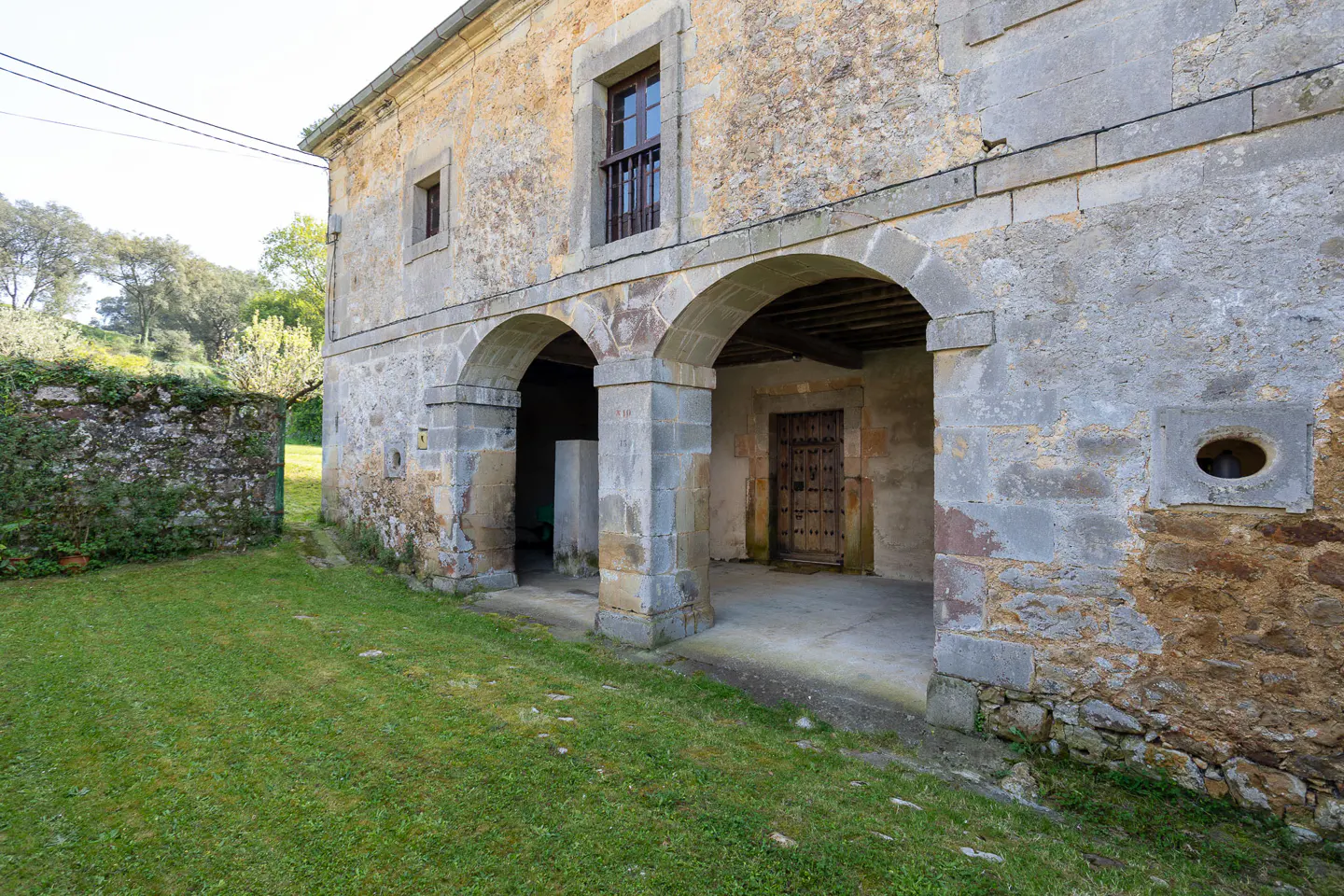Exterior view of a stone house with arched openings, a wooden door, and a green lawn.