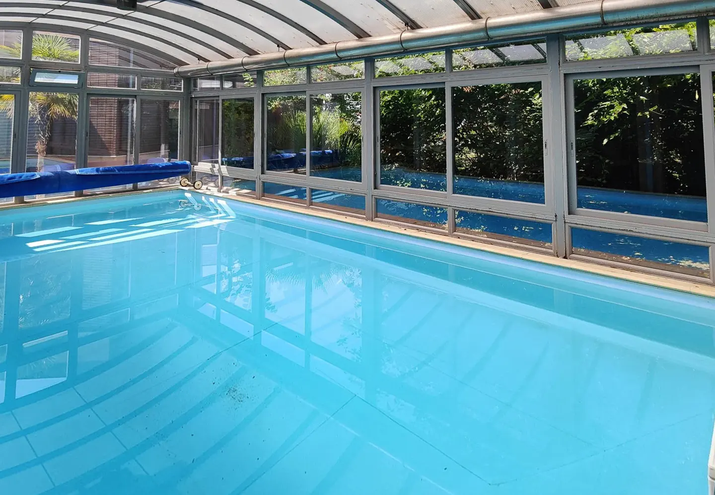 Indoor swimming pool with bright blue water, enclosed by a glass-paneled roof and walls overlooking lush green trees.