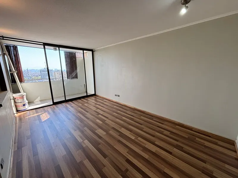 Empty room with wood floors, light gray walls, and a sliding glass door to a balcony with city views. Paint cans and ladder are visible.