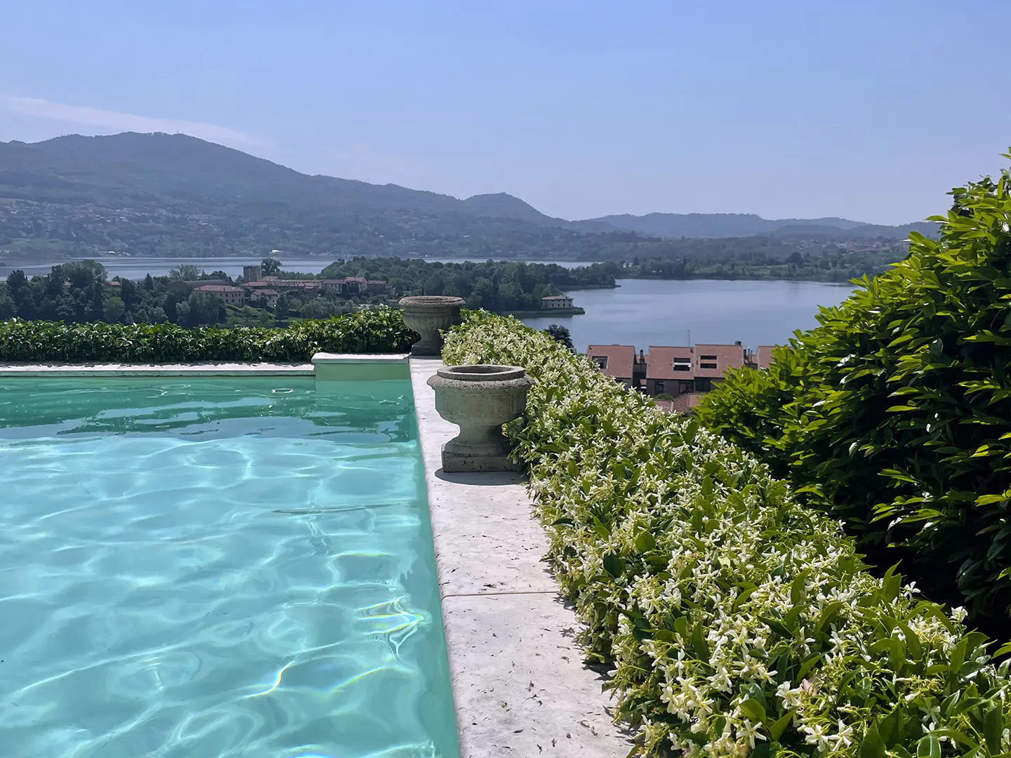 A turquoise pool overlooks a lake and mountains. Stone planters and white flowers line the pool's edge.