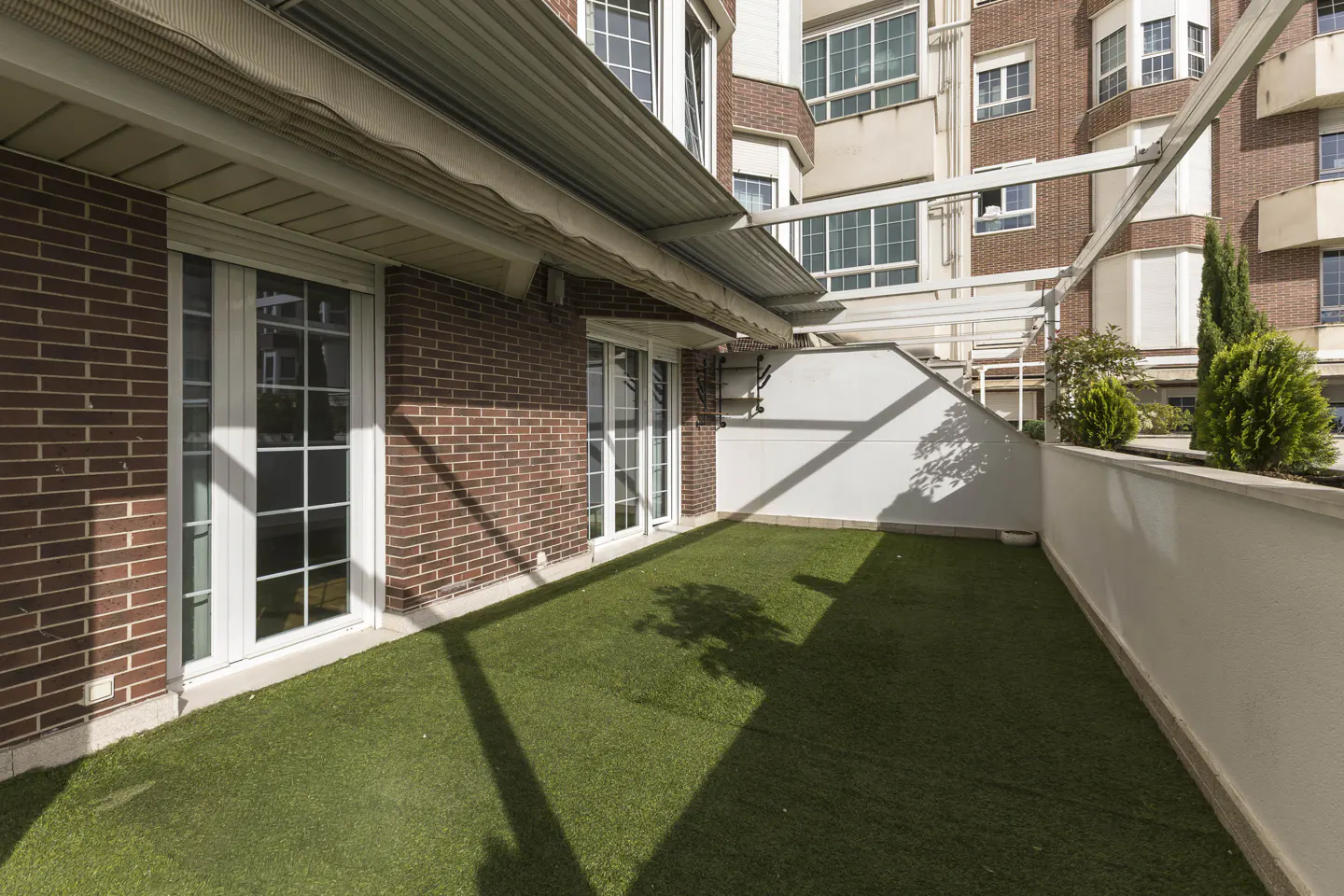 A brick patio with artificial grass, white framed windows and doors, and a white wall with greenery.