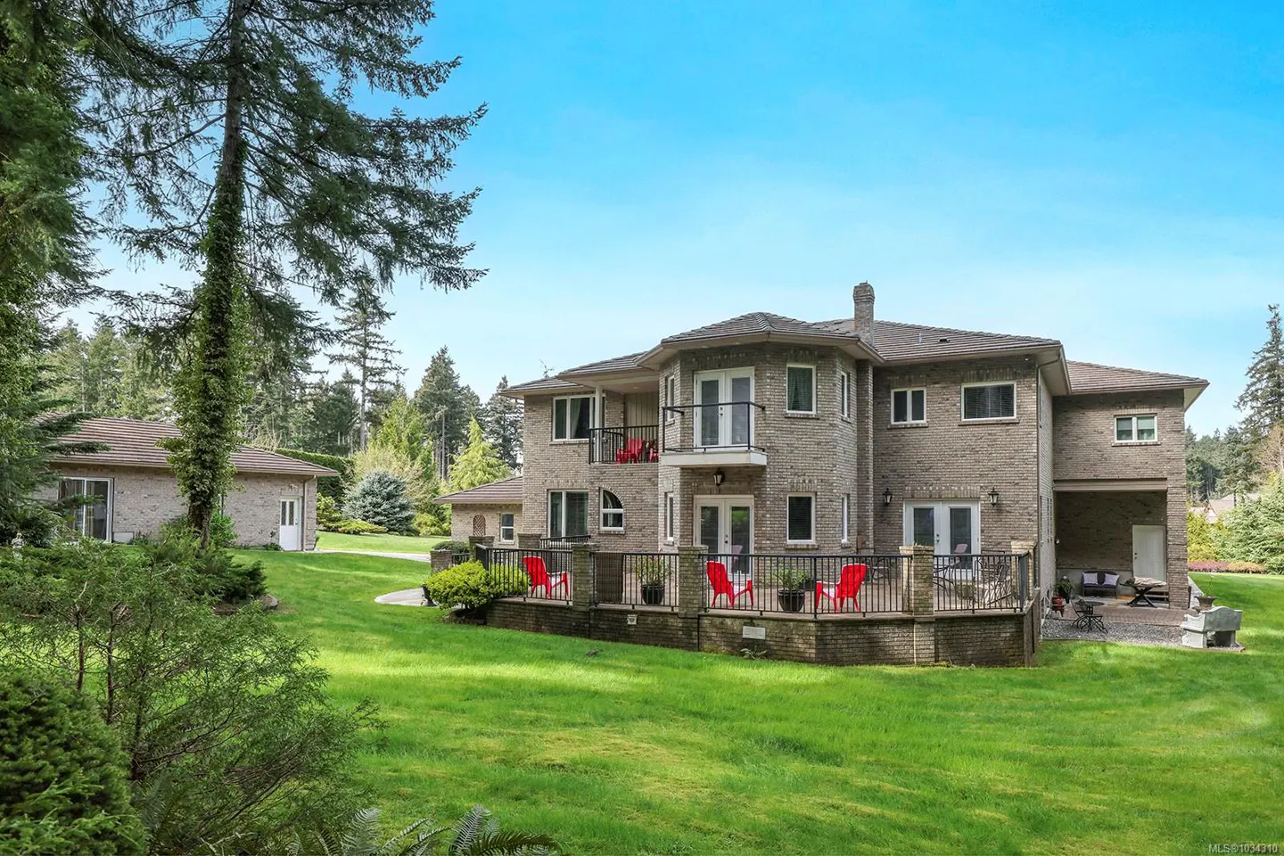 Two-story brick house with a deck and red chairs, surrounded by a green lawn and trees under a blue sky.