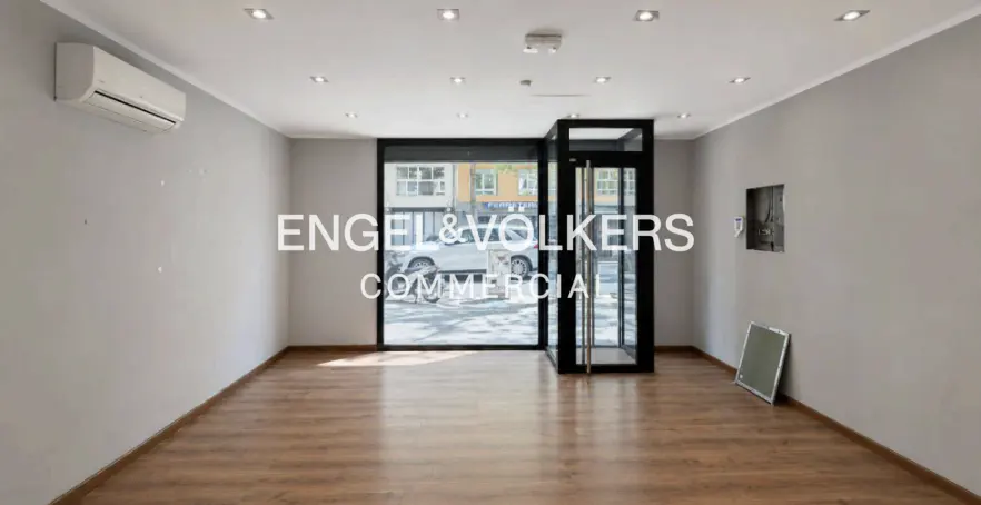 Empty commercial space with wood floors, gray walls, and recessed lighting. Glass storefront with black frame and Engel & Volkers logo.