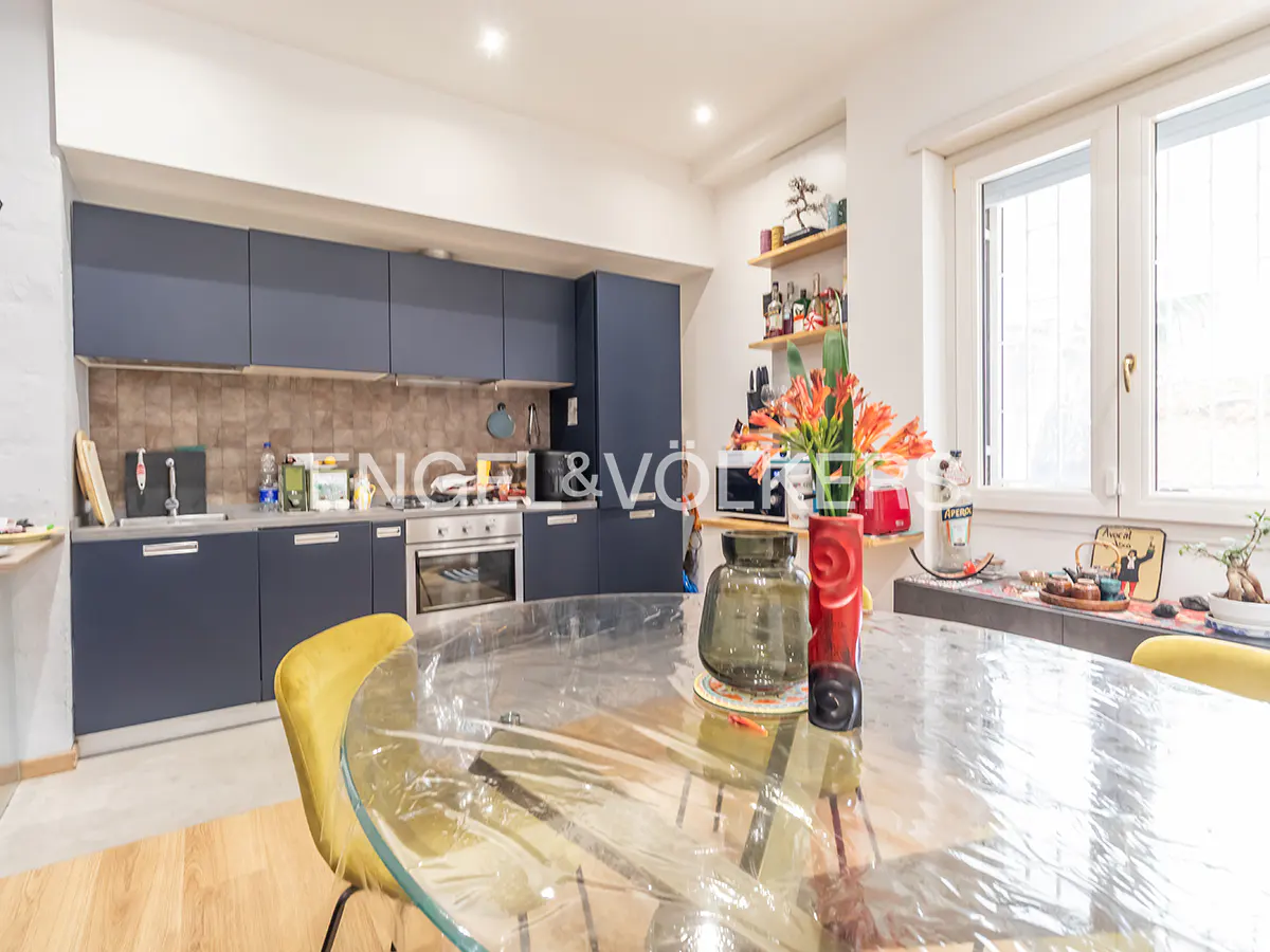 A modern kitchen with blue cabinets, stainless steel appliances, and a round glass table with yellow chairs. Orange flowers add a pop of color.