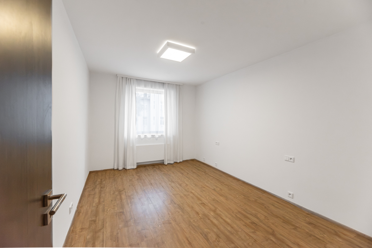 Empty room with white walls, light wood flooring, a window with white curtains, and a square ceiling light. [AI-generated text]