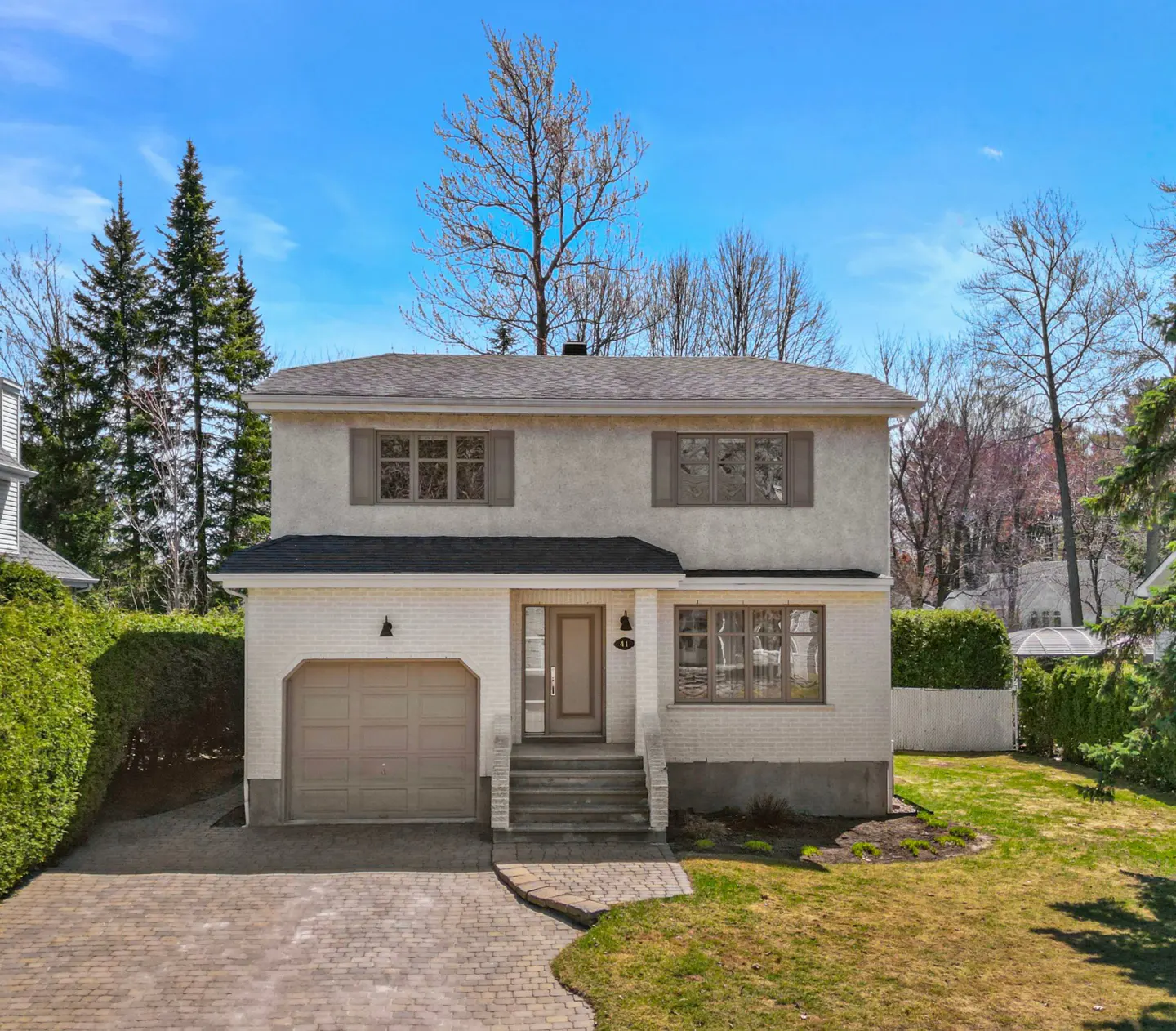Two-story house with a beige garage door, brick driveway, and green lawn under a blue sky.