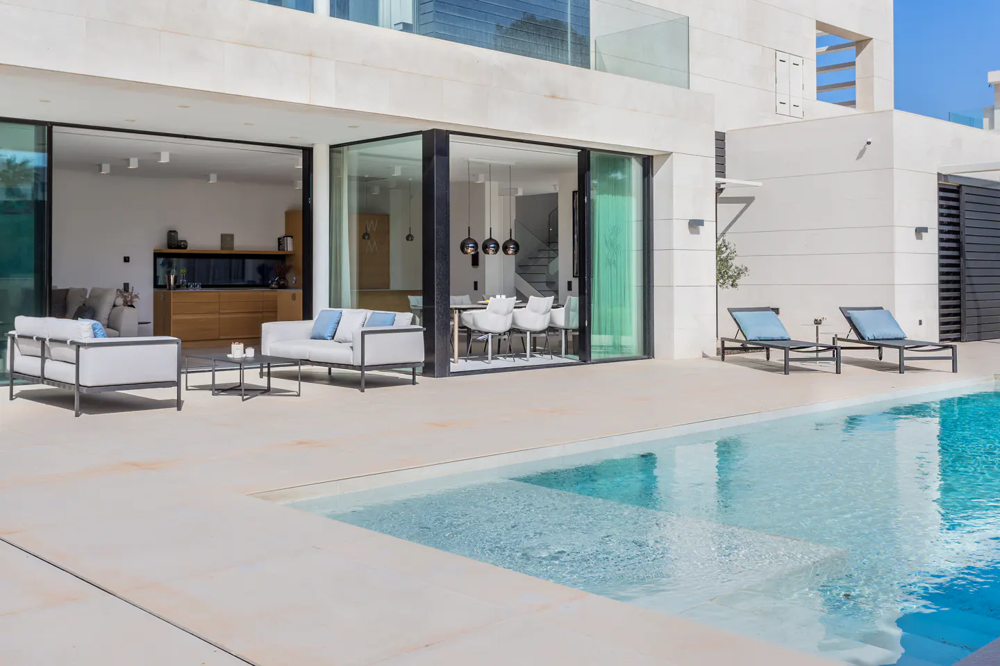 Luxury home exterior with a pool. Patio furniture includes a sofa, chairs, and loungers. Large glass doors open to the interior.