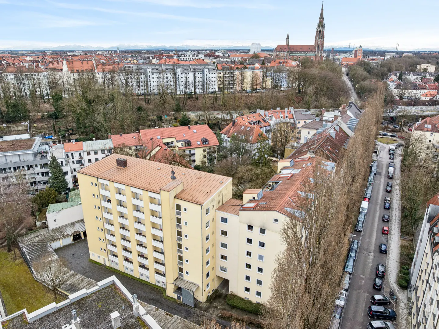 Aerial view of a yellow apartment building with a red-tiled roof, a tree-lined street with parked cars, and a cityscape in the background.