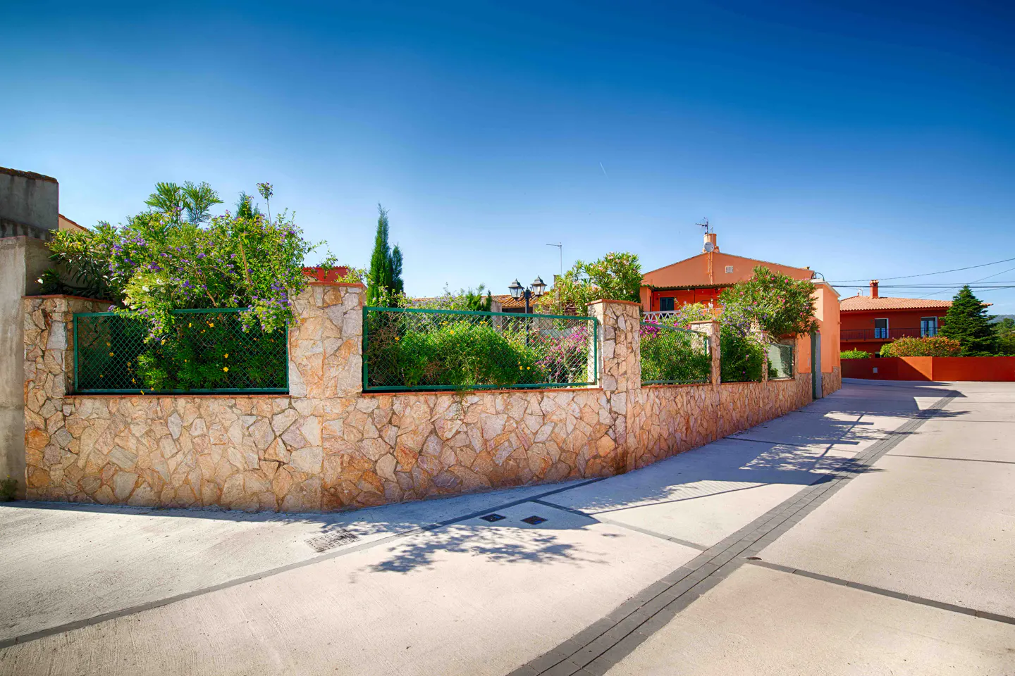 A stone wall with green fencing and foliage lines a street under a clear blue sky. Orange houses are visible behind the wall.