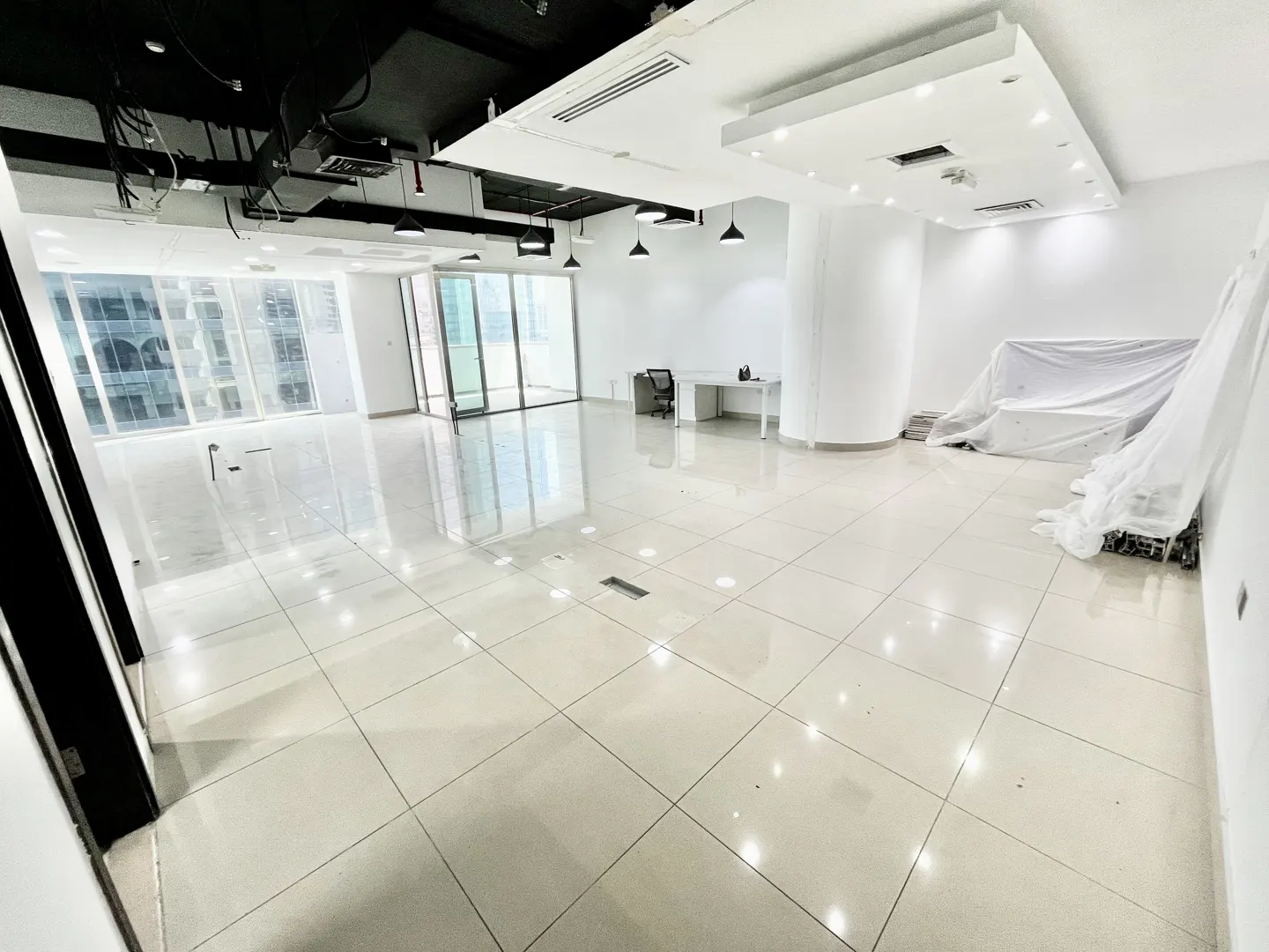 Bright, empty office space with white tile floors, large windows, and a black ceiling with hanging lights. A desk and covered furniture are visible.