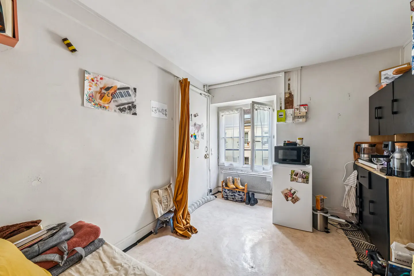 A small studio apartment with a bed, white walls, and a window. A mini fridge and microwave are near a black cabinet.