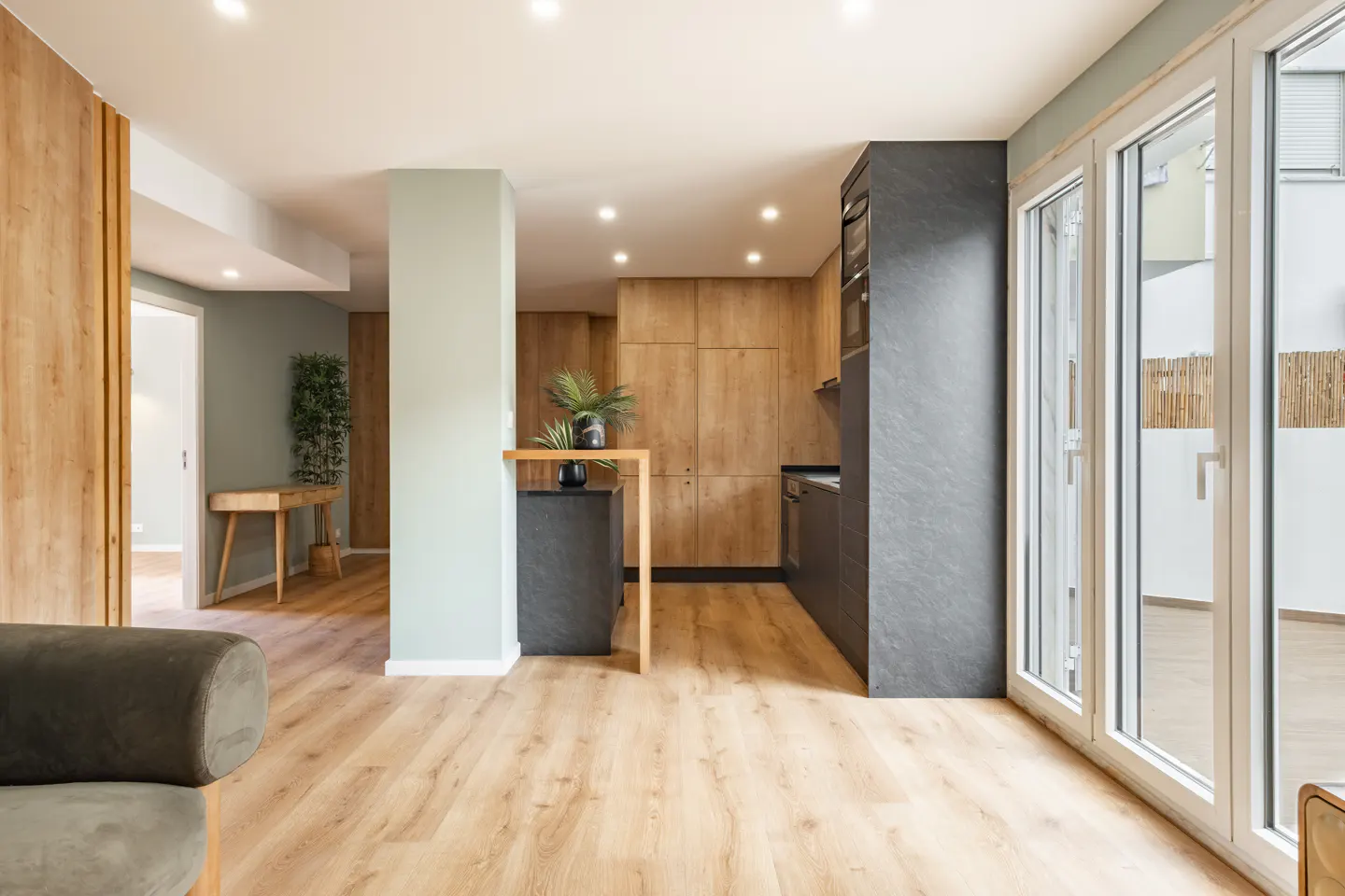 A modern apartment interior with wood floors, a gray kitchen, and large windows. A green accent wall adds a touch of color.
