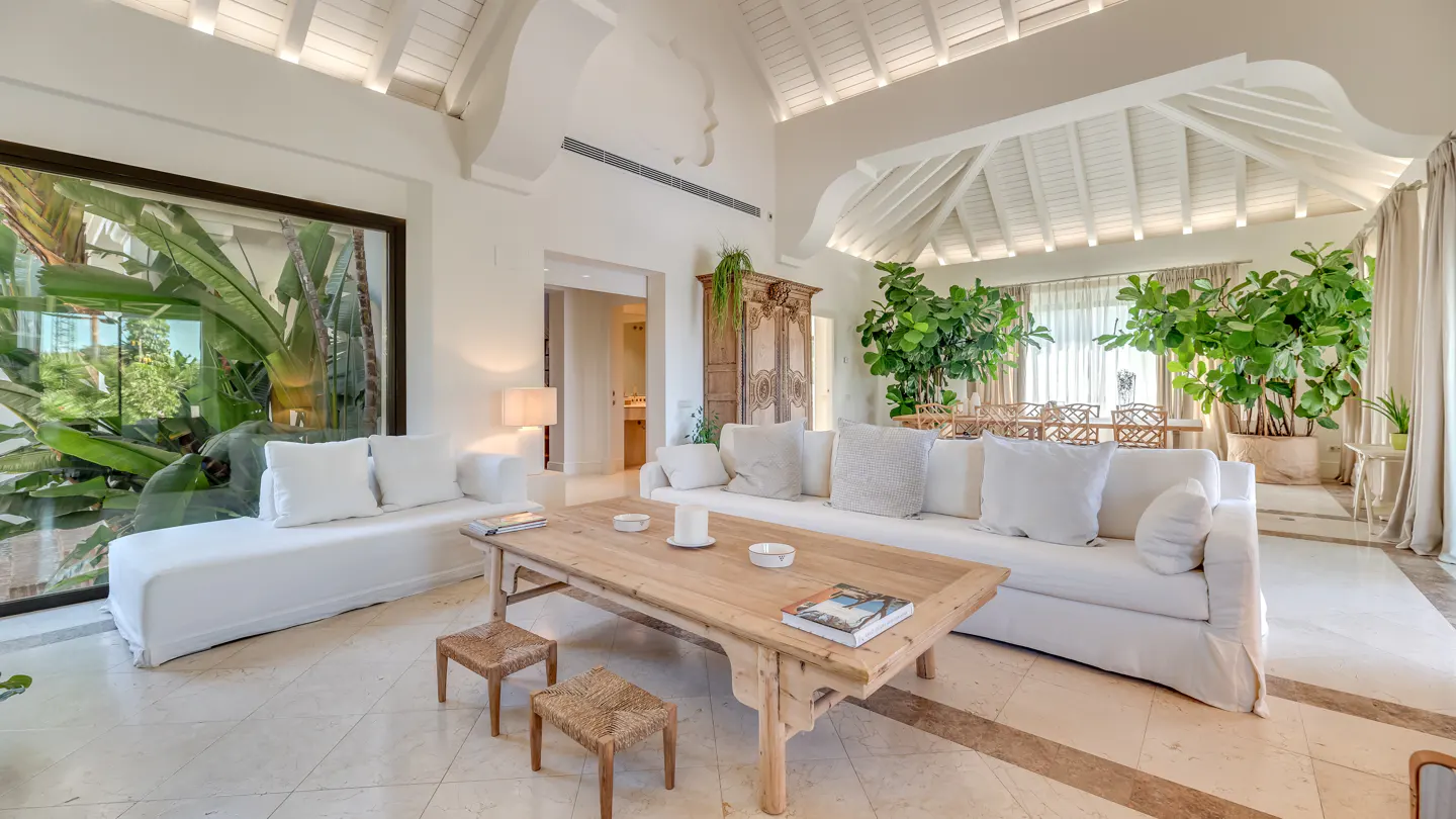 Bright living room with white sofas, a wooden table, and large windows with tropical plants. High ceilings and marble floors add to the luxurious feel.