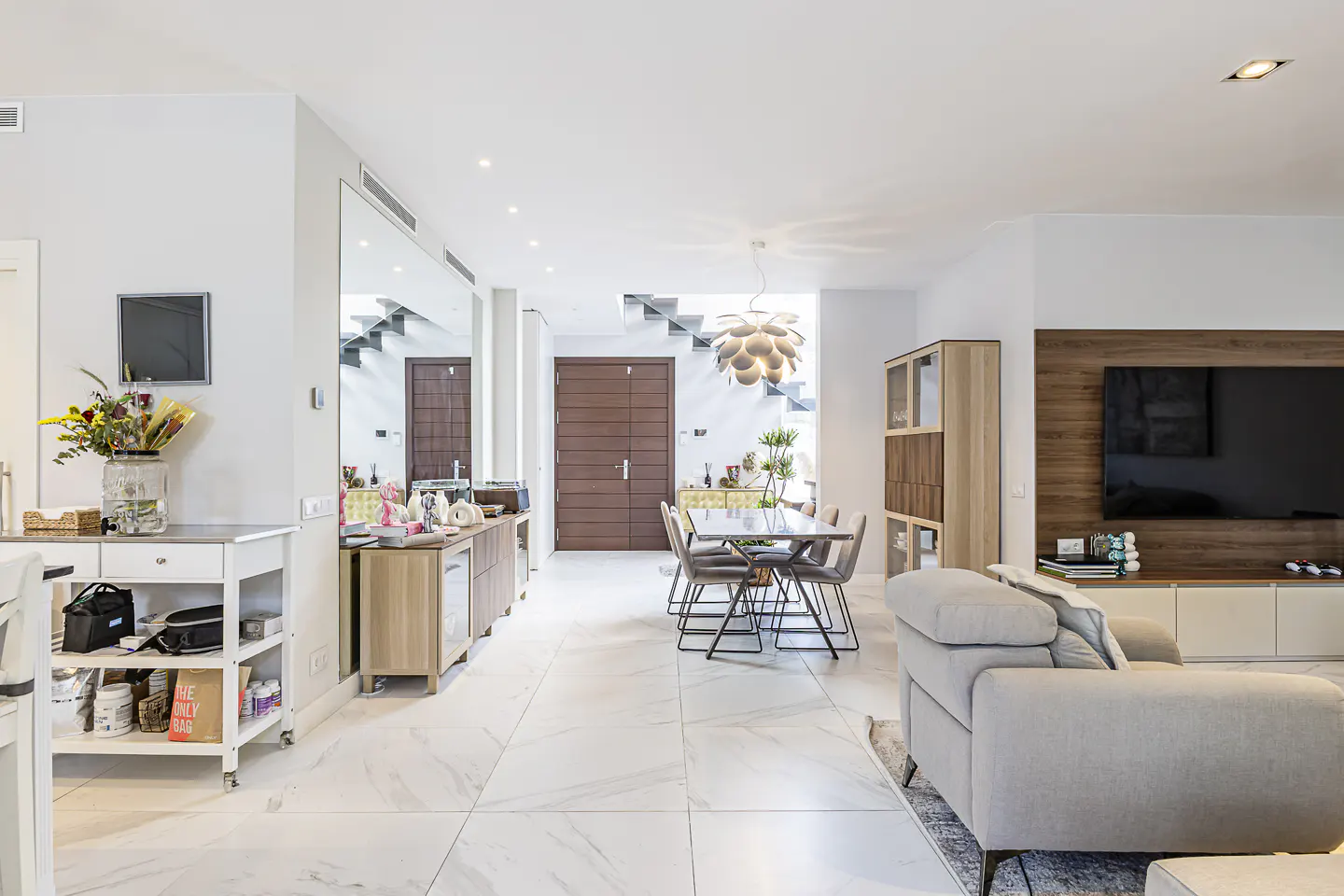 Bright, modern open-plan living space with white marble floors, dining table, and gray sofa. A large TV is mounted on a wood-paneled wall.