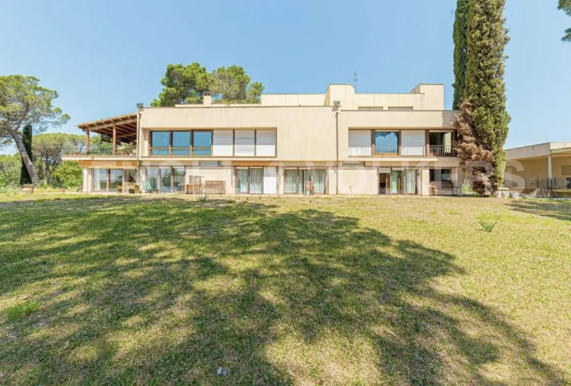 Beige two-story house with large windows and a covered balcony, surrounded by a green lawn and trees under a clear blue sky.