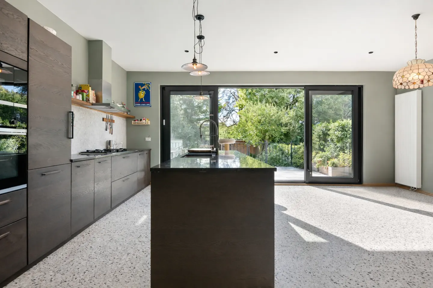 Modern kitchen with gray cabinets, black island, and terrazzo floor. Sliding glass doors open to a green backyard.