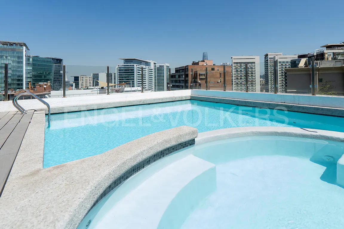 Rooftop pool and hot tub with city skyline view. Blue water, white stone, and glass railings.