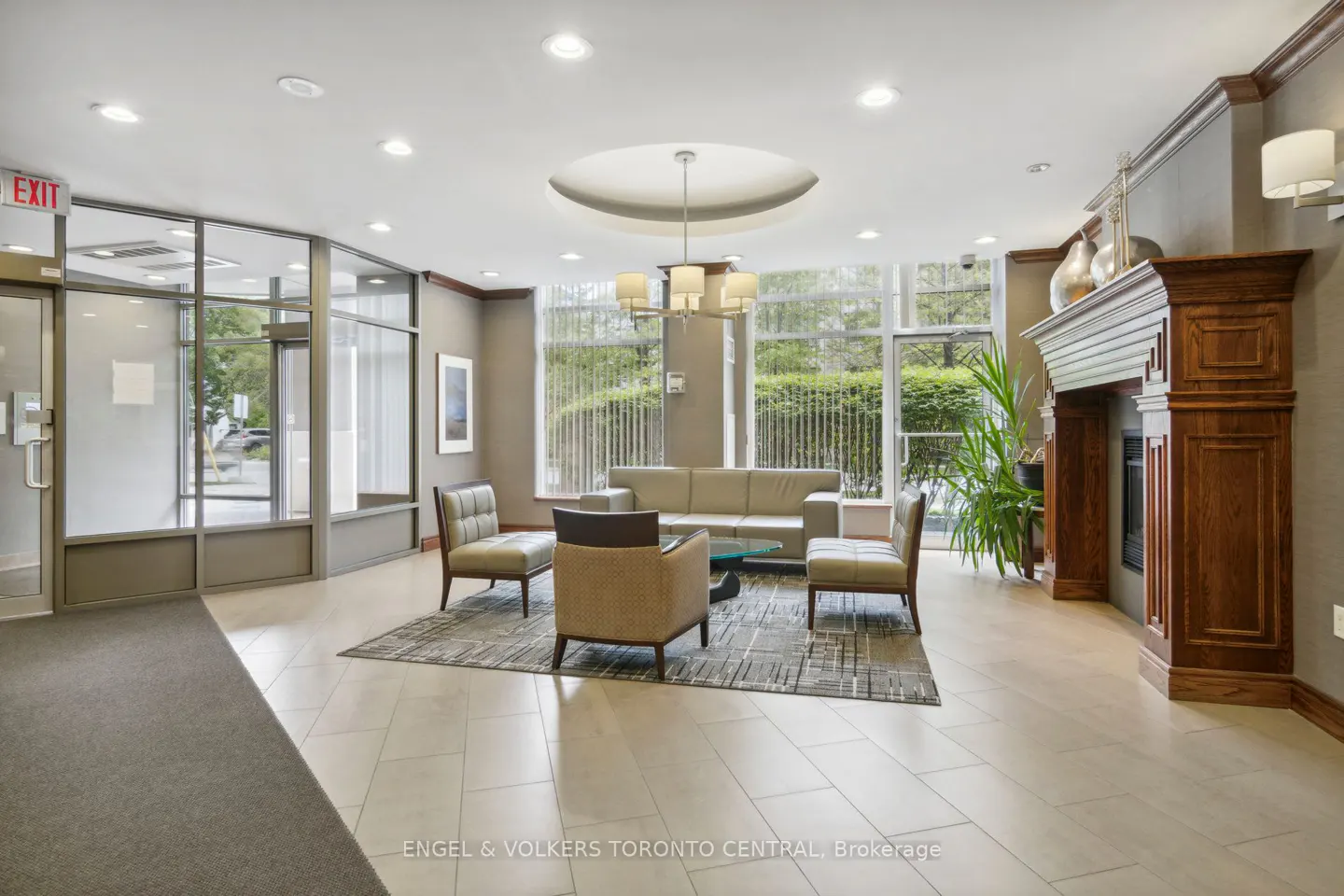 Condo lobby with seating area, fireplace, and large windows. Beige sofa and chairs sit on a patterned rug. An exit sign is visible near the entrance.