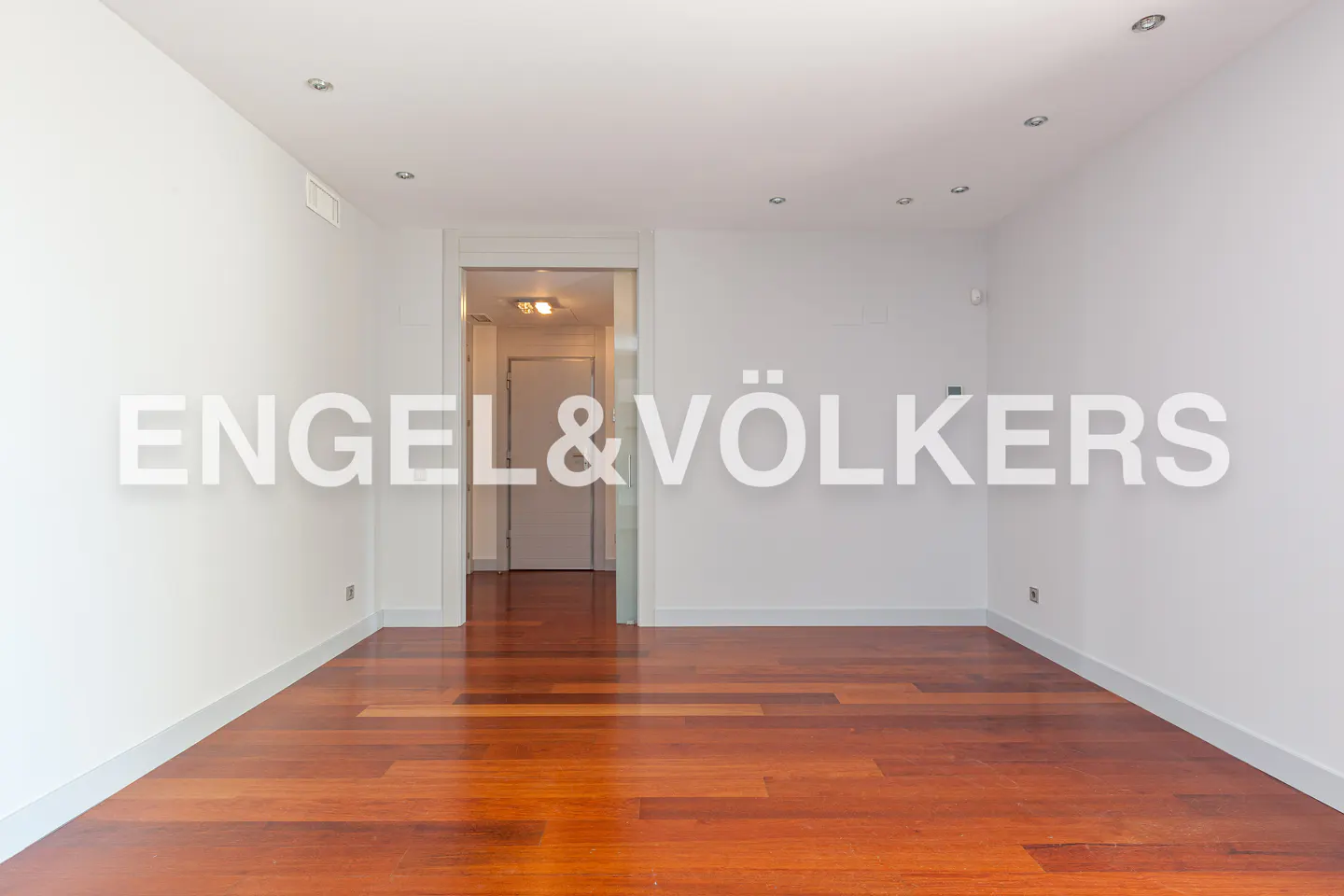Empty room with white walls, recessed lighting, and a dark wood floor. A doorway leads to a hallway. Engel & Völkers logo overlay.