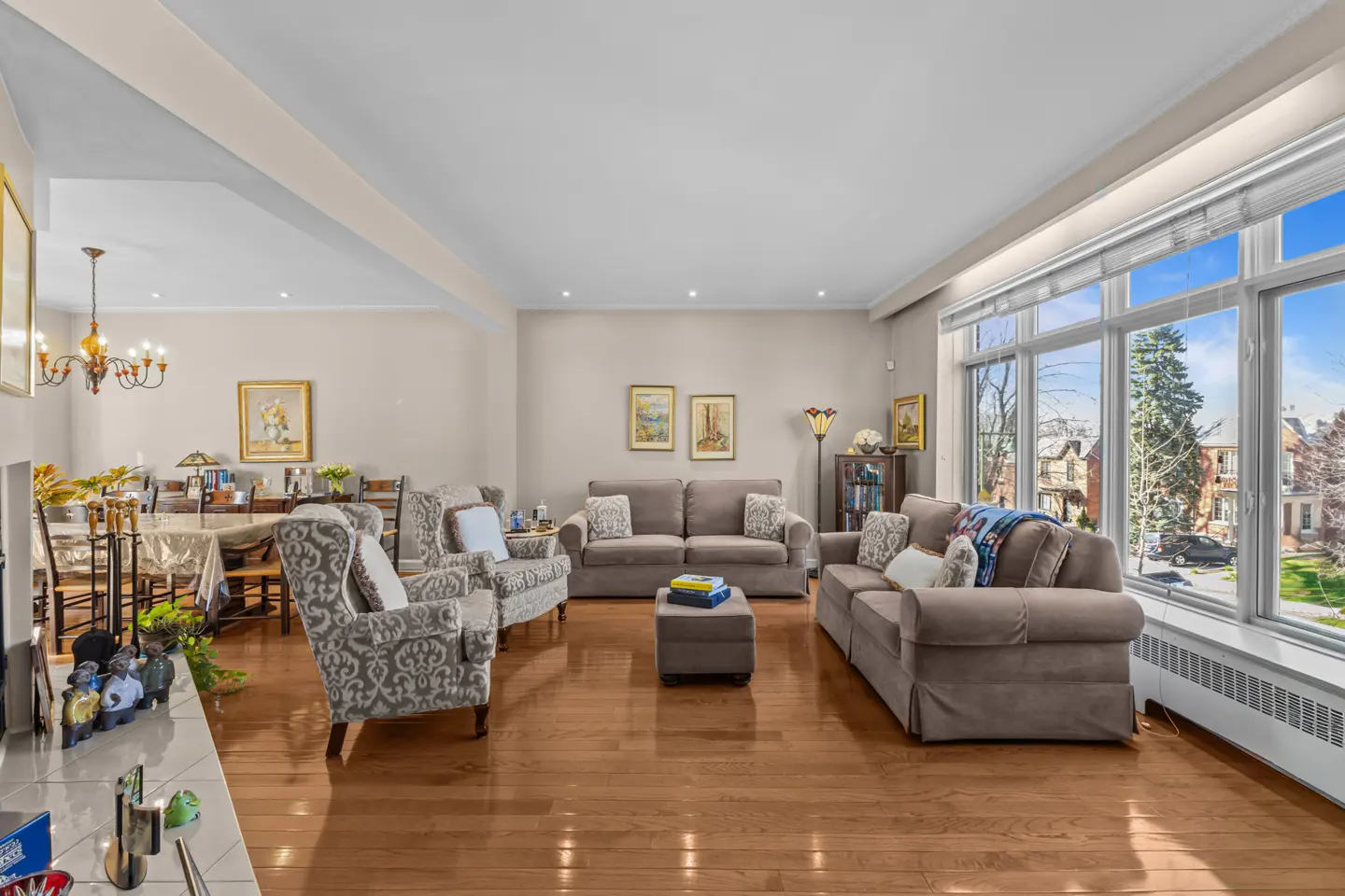 Bright living room with hardwood floors, beige walls, and large windows. Two sofas and patterned armchairs face each other. Dining area visible in the background.