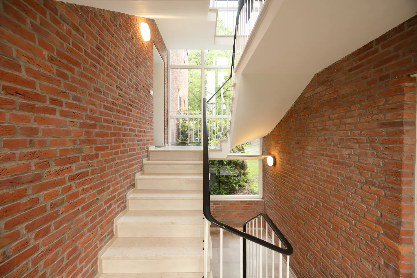 Modern staircase with light stone steps, black railings, and exposed red brick walls, illuminated by wall-mounted lights.