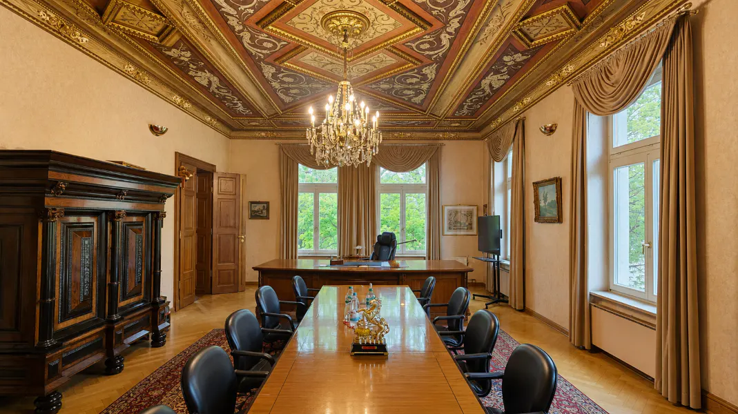 Ornate meeting room with a long wooden table, black chairs, and a crystal chandelier. Windows with beige curtains overlook greenery.