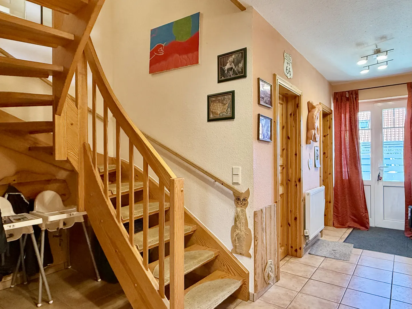 Interior view of a home's entryway with a wooden staircase, tiled floor, and peach-colored walls adorned with art. A door with red curtains is visible.
