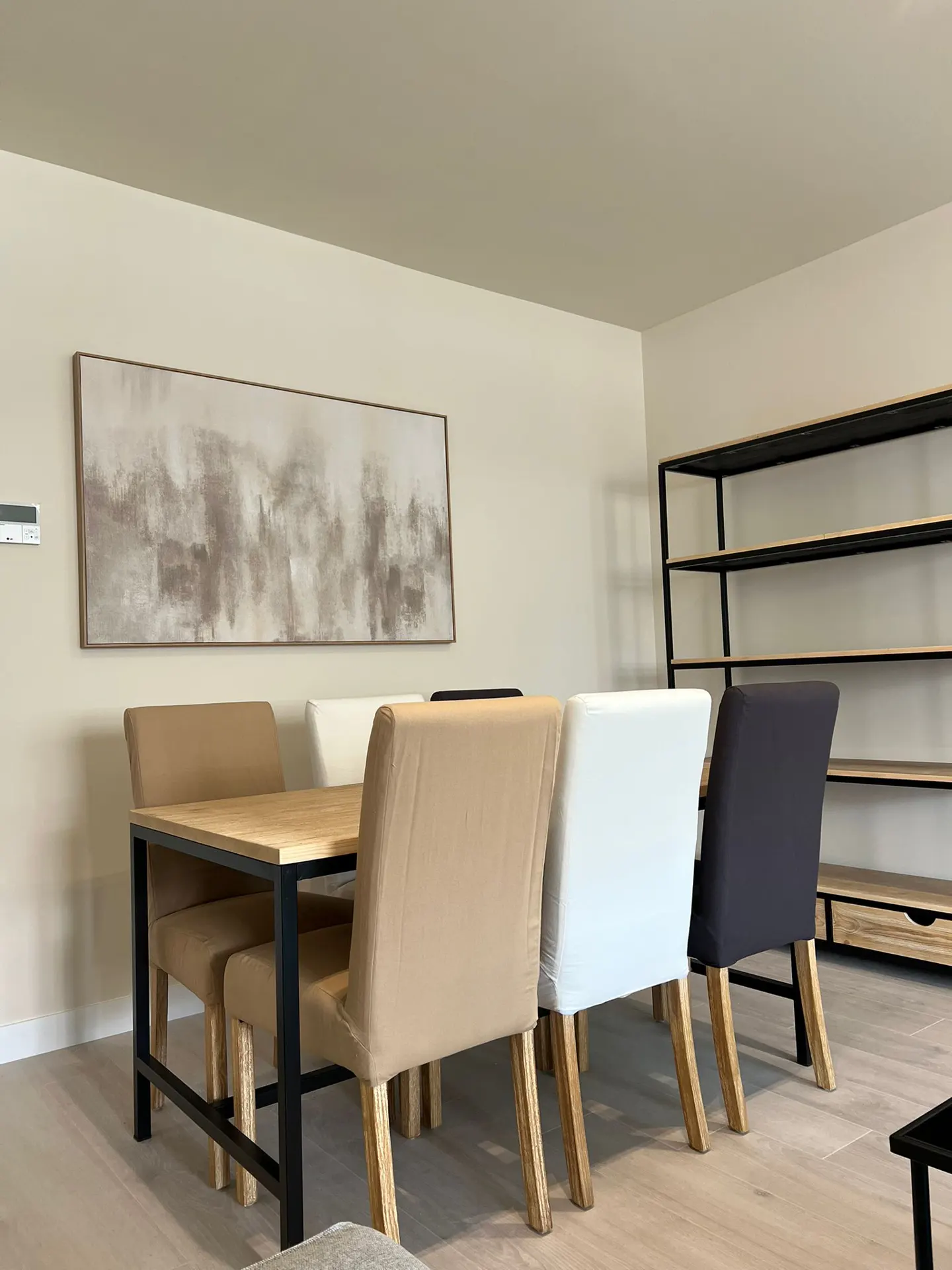 A dining room with a wooden table, black metal frame, and chairs in tan, white, and black. An abstract painting hangs on the wall.