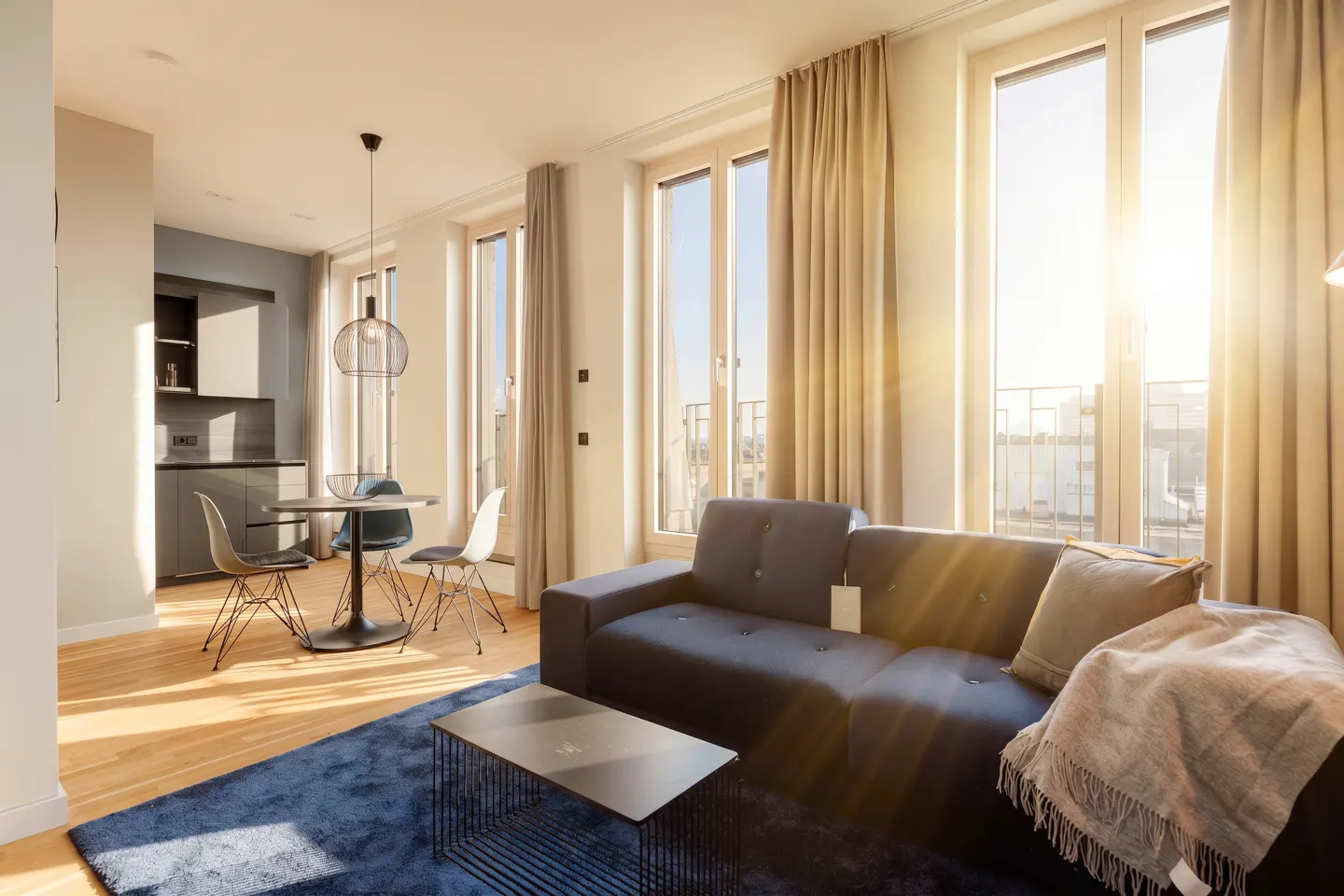 Bright, modern apartment interior with a blue sofa, rug, and a dining area near a gray kitchen. Sunlight streams through tall windows.