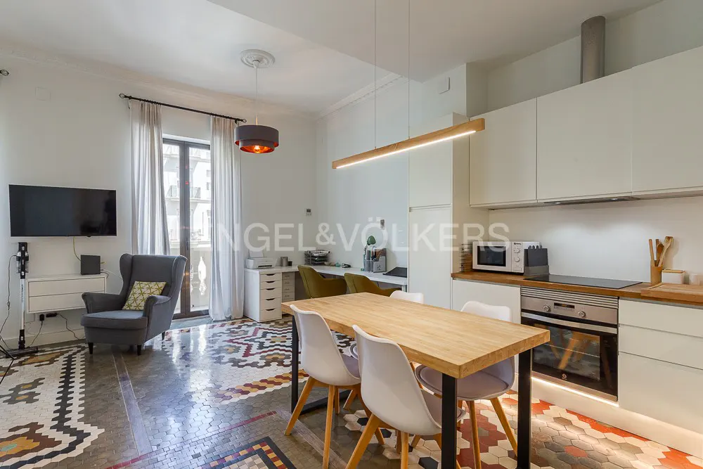 Open-concept living space with a kitchen, dining table, and lounge area. The floor has a colorful geometric pattern. White cabinets and a wooden table.