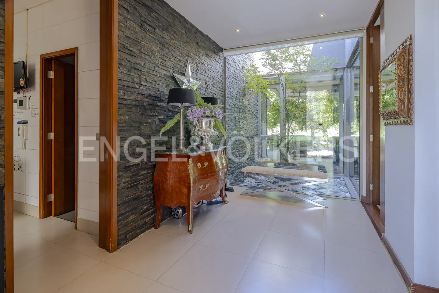 Interior view of a home's entryway with stone wall, wood dresser, and large glass window overlooking a garden.