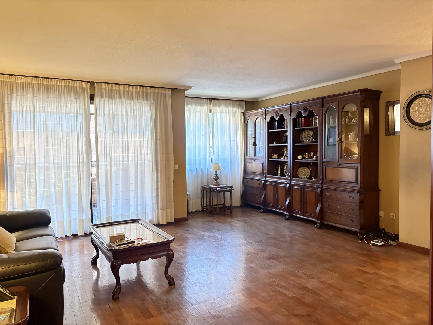 A living room with hardwood floors, a dark brown sofa, a glass coffee table, and a large dark wood bookcase. Light streams in through sheer white curtains.