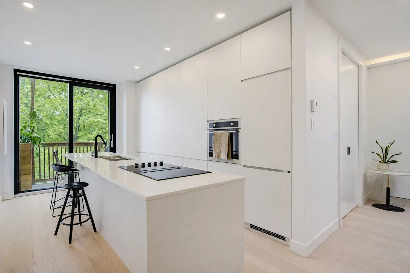 Bright, modern kitchen with white cabinets, island with black stovetop, and black stools. Sliding glass doors open to a balcony with green trees.