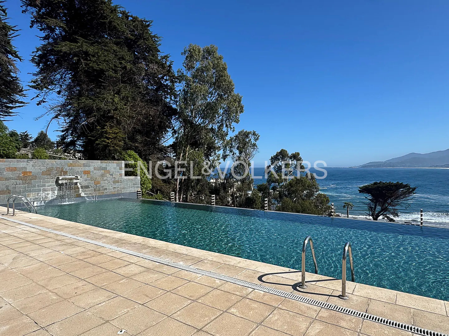 Infinity pool overlooking the ocean on a sunny day. Trees and mountains are in the background.