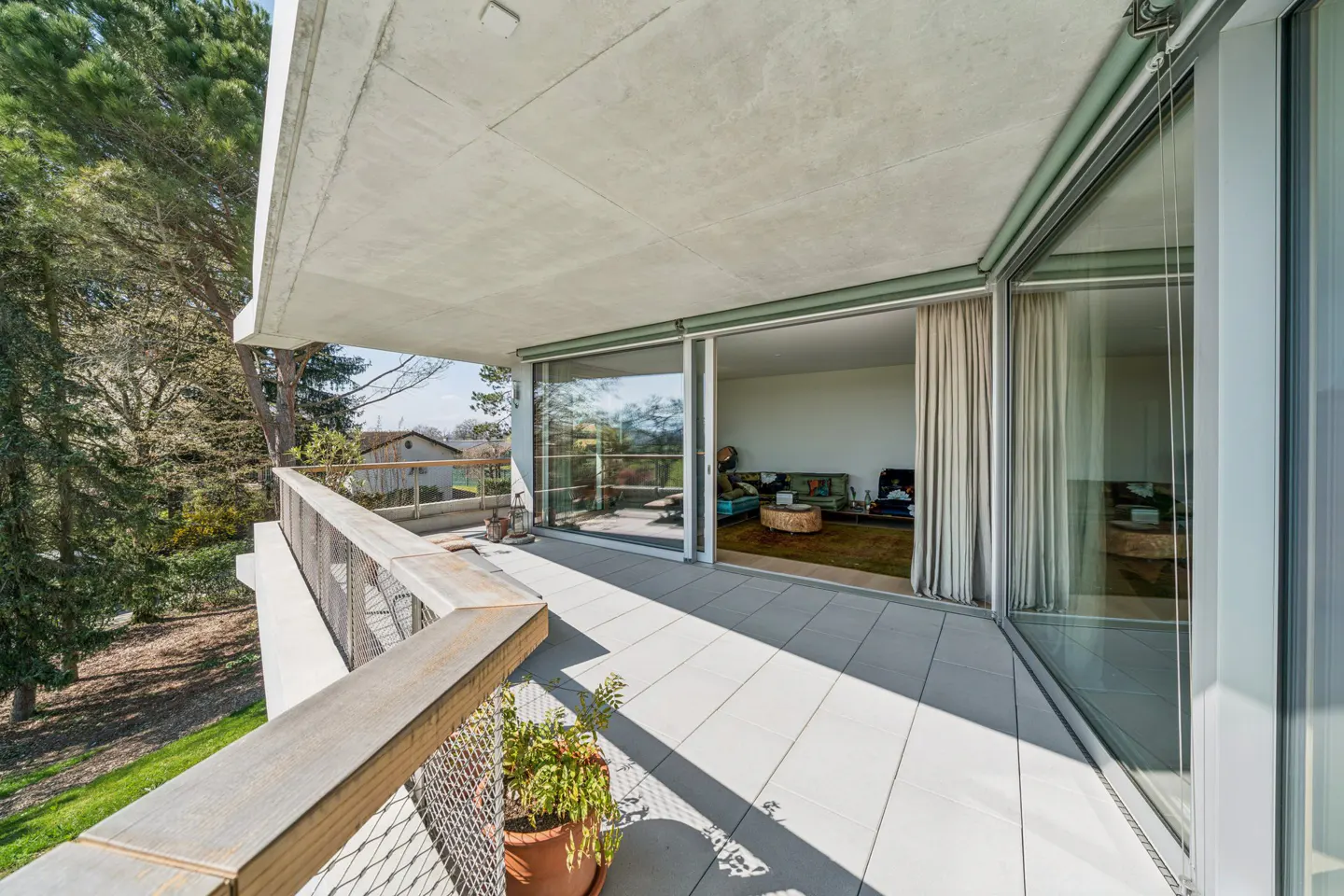 Wide balcony with gray tiles, metal railing, and potted plant. Sliding glass doors open to a living room with a patterned sofa and round coffee table. Trees in the background.