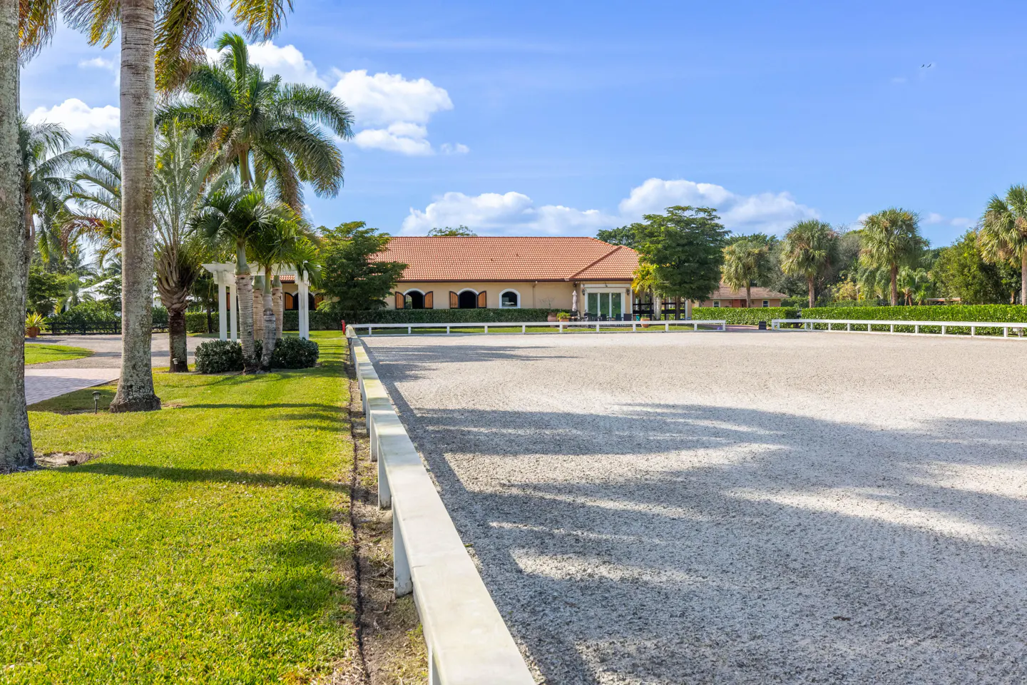 Outdoor horse riding arena with white fence, sandy ground, and palm trees. A building with a red tile roof is in the background.