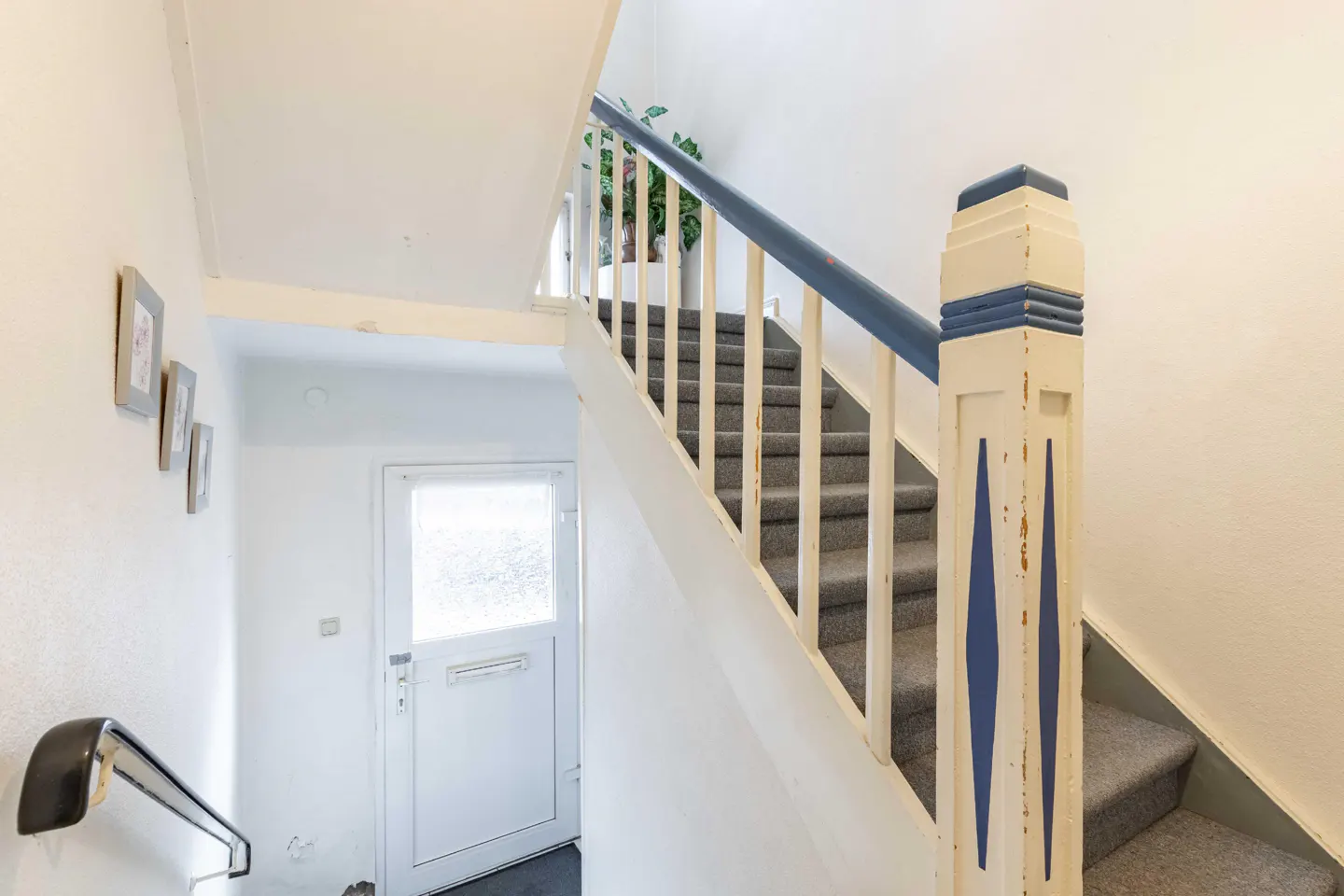 Interior view of a staircase with gray carpet, white walls, and a blue handrail. A white door is visible at the bottom of the stairs.