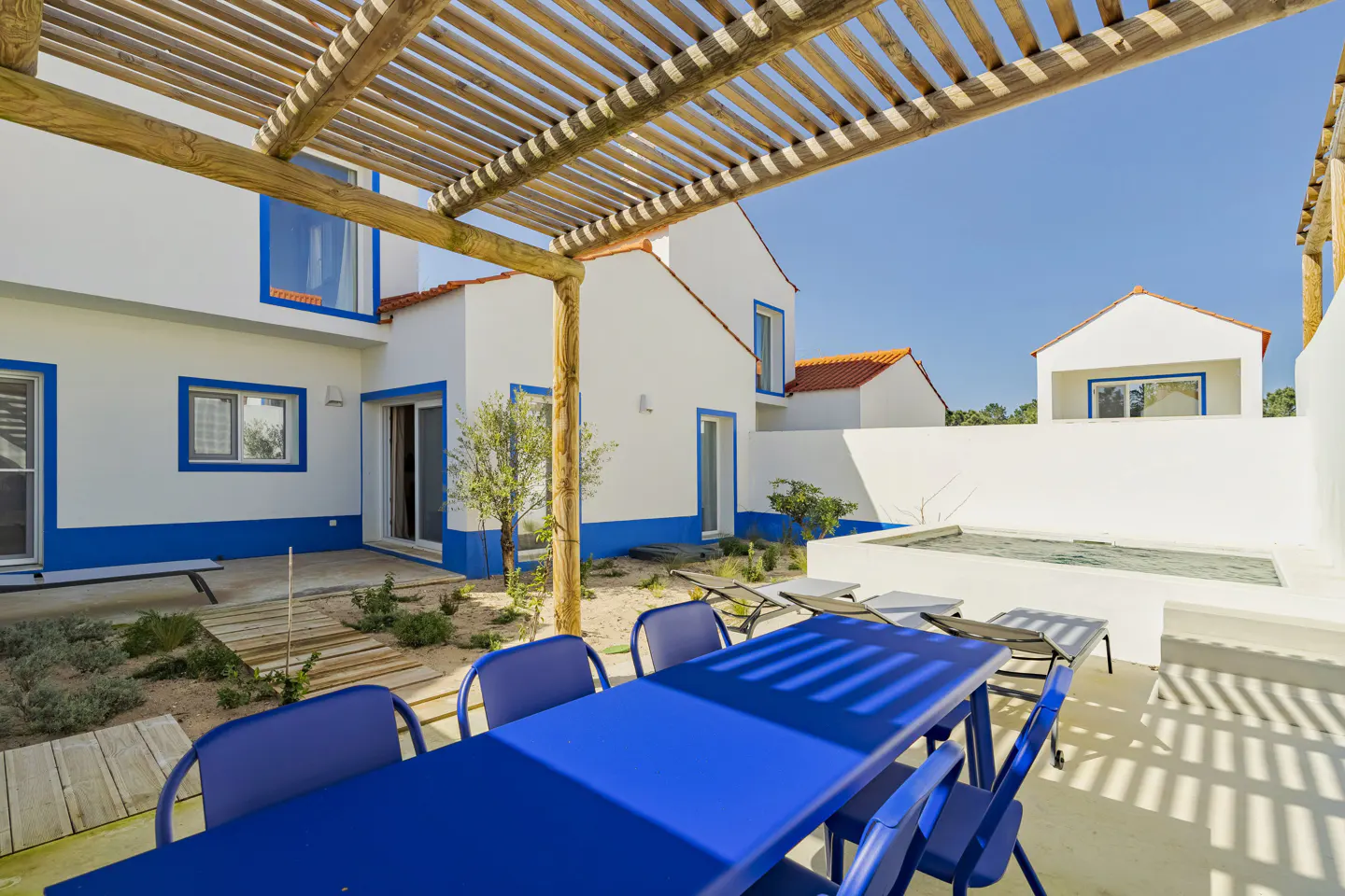 Outdoor patio with a blue table and chairs under a wooden pergola. White buildings with blue trim and orange roofs surround the patio.