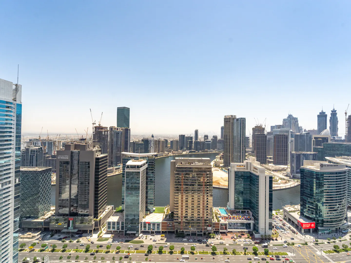 High-angle view of a modern Dubai skyline with glass skyscrapers, a central canal, and a busy road under a clear blue sky.