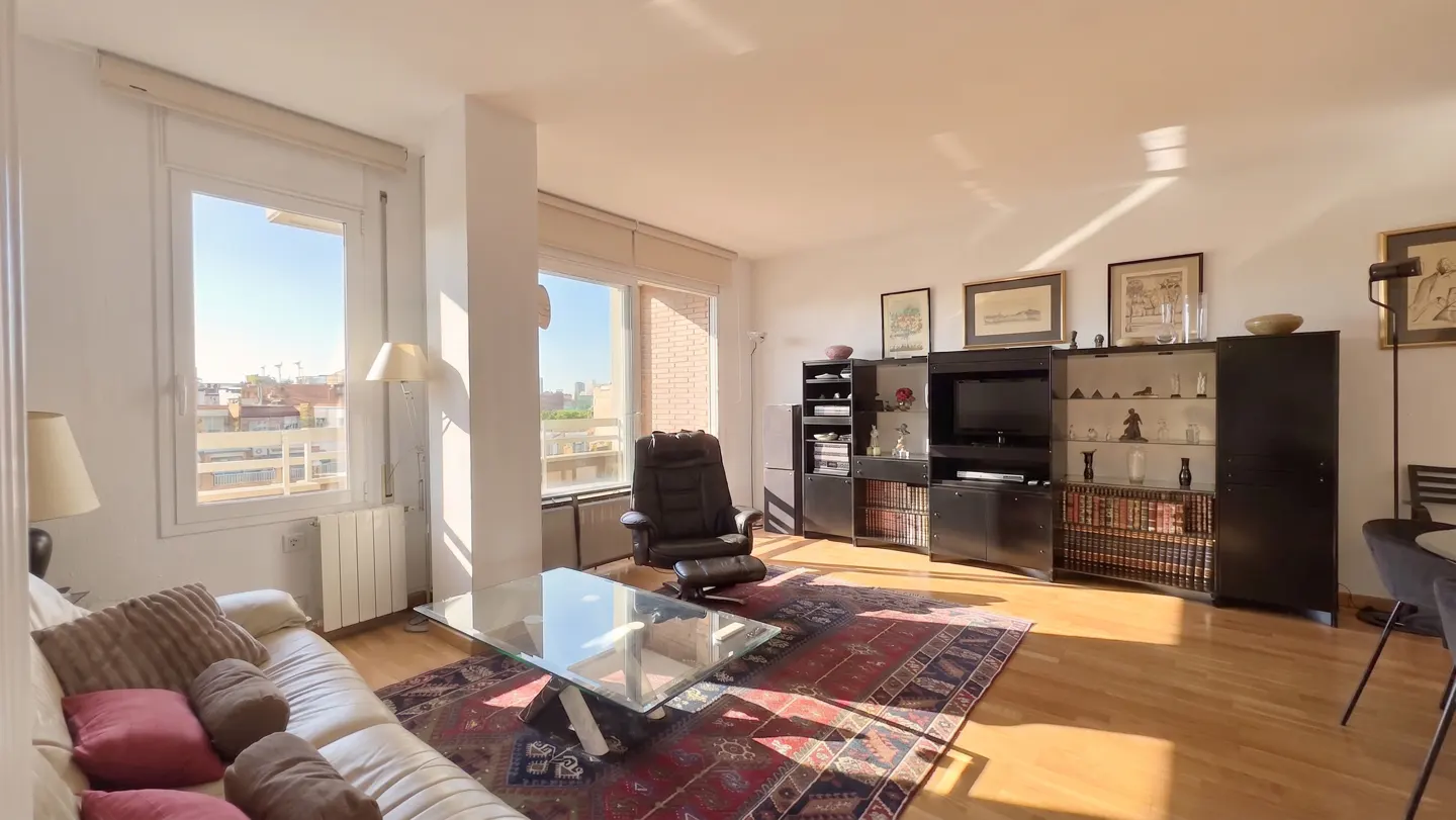 Sunlit living room with a white sofa, glass table, black shelves, and a large window overlooking a cityscape.
