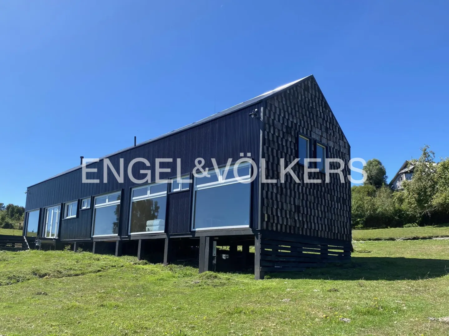 Black modern house with large windows on a green lawn under a blue sky. Engel & Völkers logo overlay.