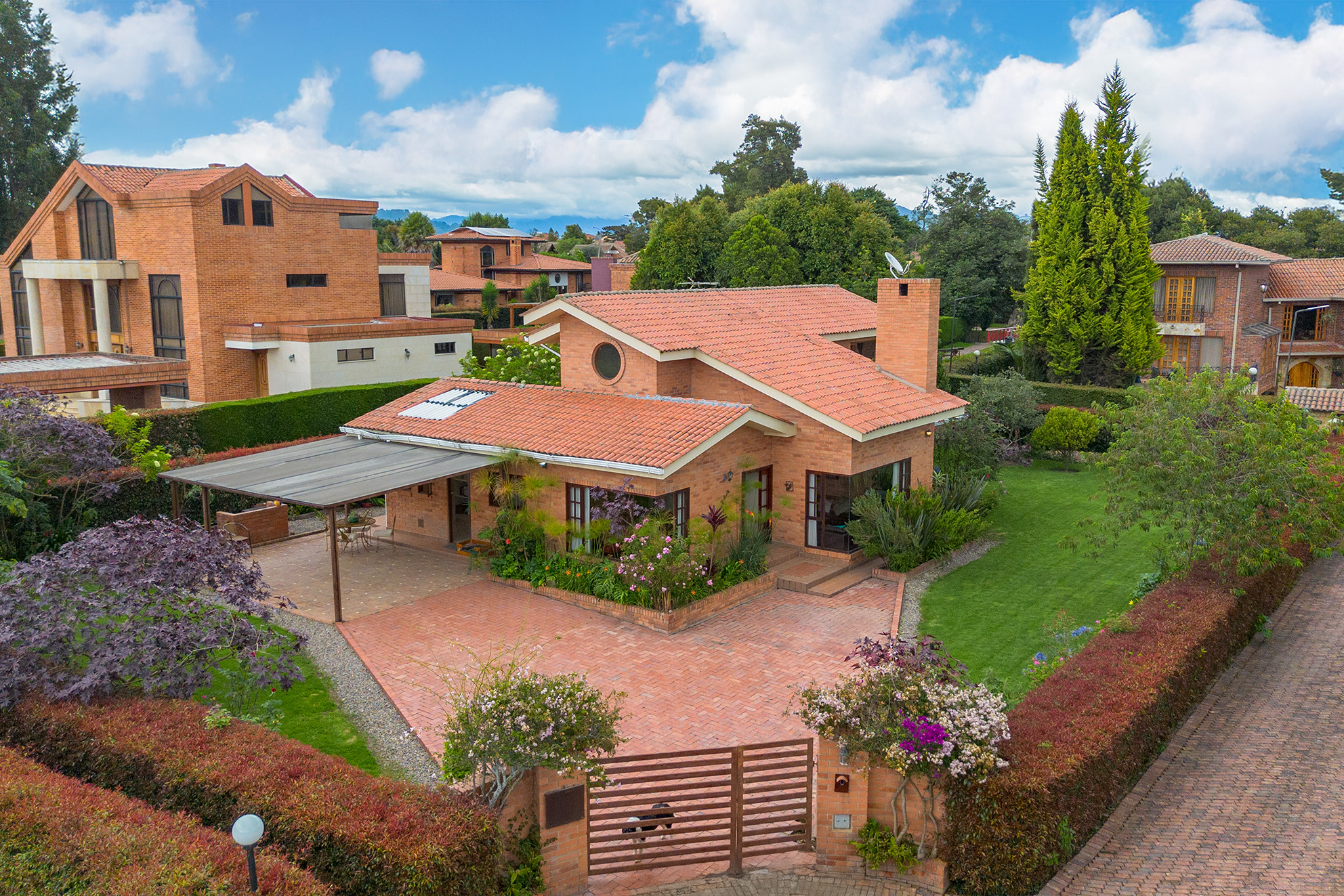 Brick house with a red tile roof, surrounded by green lawns and hedges, under a blue sky with white clouds. A metal gate leads to the brick driveway.