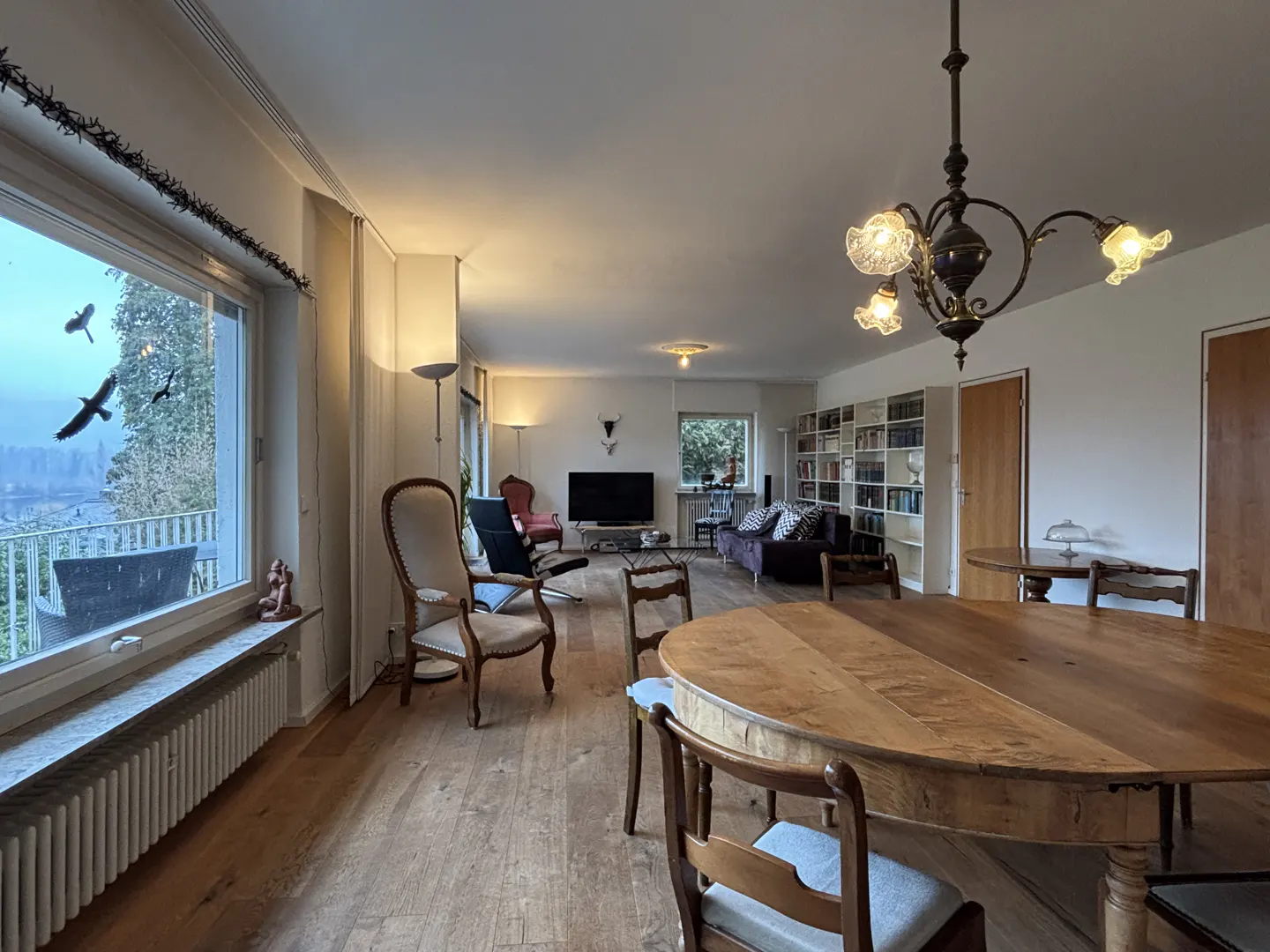 A spacious living room with hardwood floors, a large window with birds flying outside, and a dining table with chairs.