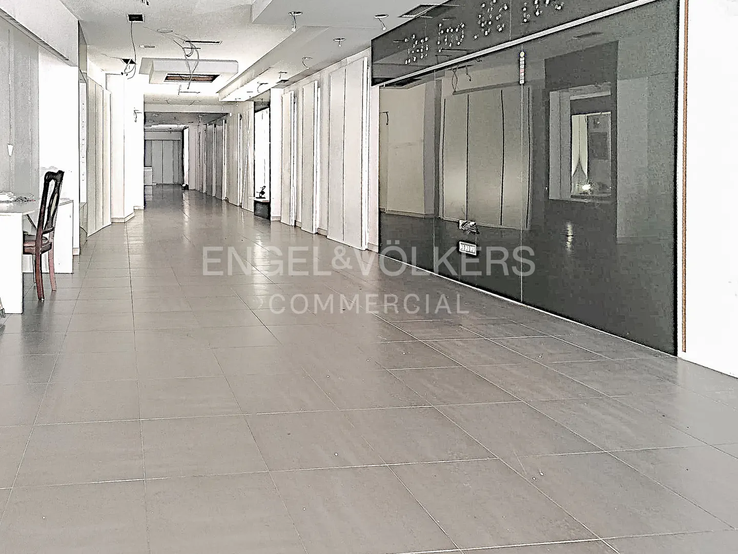 Empty commercial space with gray tile floor, white walls, and black glass facade. Engel & Völkers Commercial logo visible.