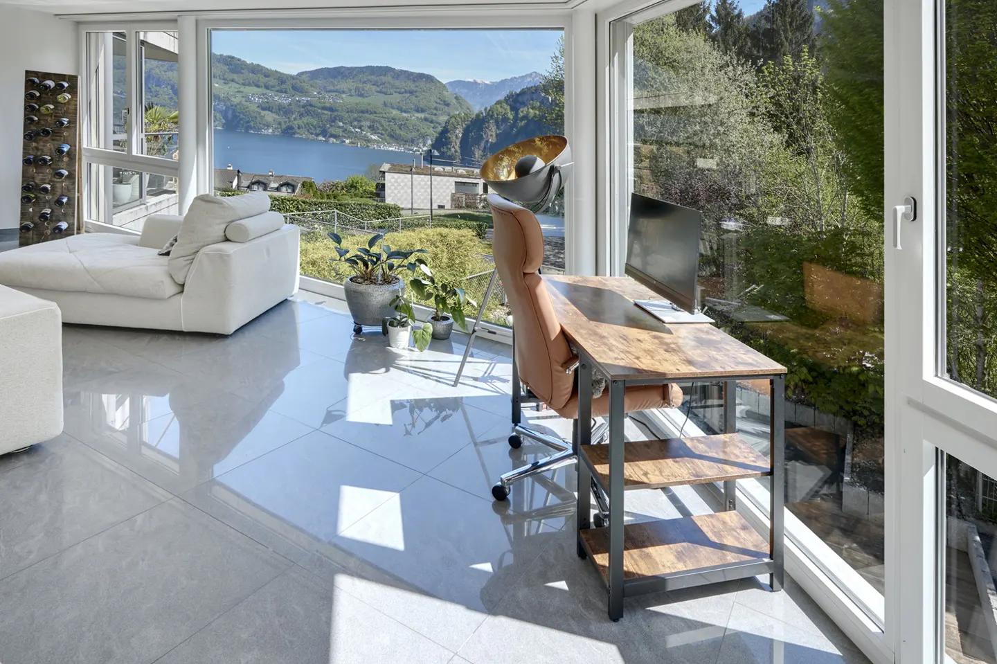 Modern home office with a desk, tan chair, and computer overlooking a scenic lake and mountain view through large windows.