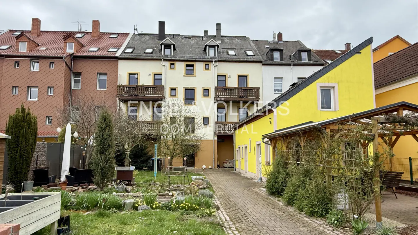 Exterior view of multi-story buildings with balconies, a yellow house, and a garden with a brick path.